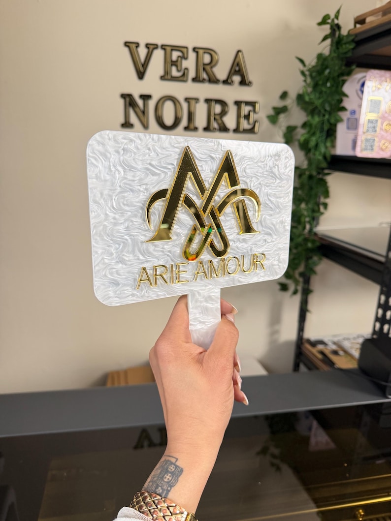 May include: A white, rectangular sign with a handle, featuring gold lettering that reads "ARIE AMOUR" and a gold emblem. The sign has a textured, marbled appearance. The words "VERA NOIRE" are displayed in gold letters on the wall behind the sign.