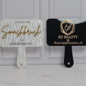 May include: Two acrylic makeup artist hand mirrors. One is white with gold text that reads "Makeup by Smashbrush" and the Instagram handle. The other is black with gold text that reads "ED BEAUTY" and the Instagram handle.
