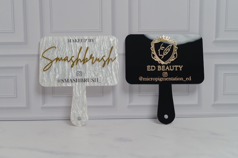 May include: Two acrylic makeup artist hand mirrors. One is white with gold text that reads "Makeup by Smashbrush" and the Instagram handle. The other is black with gold text that reads "ED BEAUTY" and the Instagram handle.