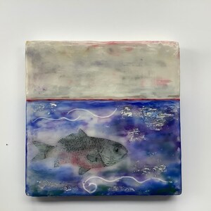 May include: A square ceramic tile featuring a painted scene of a fish swimming in blue water. The top half of the tile is a light grey with a hint of pink. The bottom half is a blue and purple watercolour wash with white swirls and a single fish in the centre. The fish is grey with a pink belly and is facing to the right.