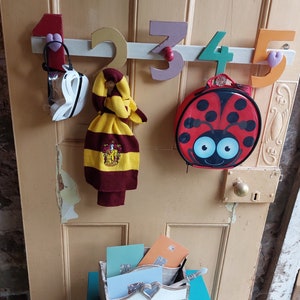 May include: A wooden coat rack with colorful wooden numbers 1 through 5. A white face mask, a yellow and maroon scarf, and a red ladybug lunch bag are hanging on the rack. A small teal blue table with a wooden box and cards on top.