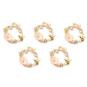 May include: Five gold-toned wreath charms, each featuring a light pink flower, clear crystals, and a small black and yellow bee. The wreaths have a floral and bee theme, perfect for jewelry making or crafting.