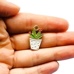 May include: A small, white enamel charm shaped like a potted plant. The pot features black polka dots. The plant has green leaves and red flowers. The charm has a gold-coloured loop at the top for attaching to jewellery.