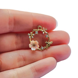 May include: A gold-colored charm with a floral wreath design. The wreath features a light pink flower, small white flowers, and a small bee. The charm is held in a hand against a white background.