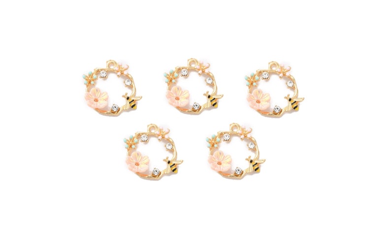 May include: Five gold-toned wreath charms, each featuring a light pink flower, clear crystals, and a small black and yellow bee. The wreaths have a floral and bee theme, perfect for jewelry making or crafting.
