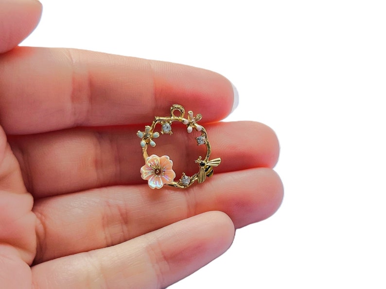 May include: A gold-colored charm with a floral wreath design. The wreath features a light pink flower, small white flowers, and a small bee. The charm is held in a hand against a white background.