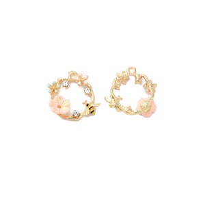 May include: Two gold-colored metal charms. One charm features a wreath design with a bee, flowers, and crystals. The other charm has a wreath design with flowers. The charms are on a white background.