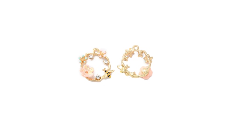 May include: Two gold-colored metal charms. One charm features a wreath design with a bee, flowers, and crystals. The other charm has a wreath design with flowers. The charms are on a white background.