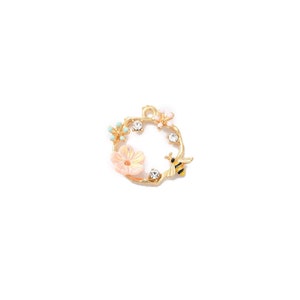 May include: A gold-colored charm with a floral and bee design. The charm features a circular wreath with a light pink flower, small blue flowers, and a yellow and black bee. Clear crystals are set around the wreath.