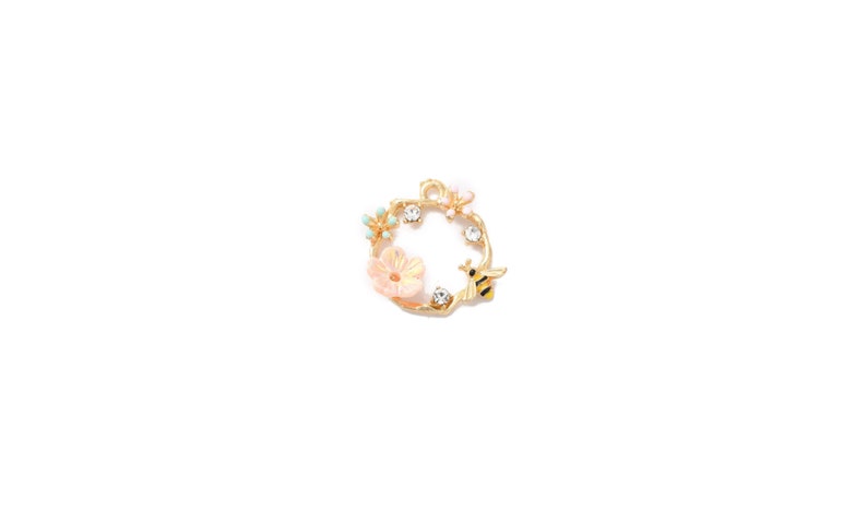 May include: A gold-colored charm with a floral and bee design. The charm features a circular wreath with a light pink flower, small blue flowers, and a yellow and black bee. Clear crystals are set around the wreath.