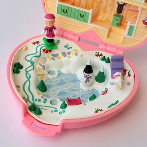 Può includere: Un playset Polly Pocket rosa vintage, aperto per rivelare una scena invernale. Il set include un mini sciatore, un pupazzo di neve e altri accessori a tema invernale. Il playset è a forma di cuore e presenta un interno dettagliato.