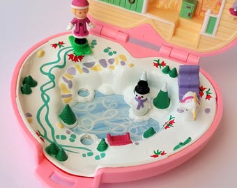 May include: A vintage pink Polly Pocket playset, open to reveal a winter scene. The set includes a miniature skier, snowman, and other winter-themed accessories. The playset is heart-shaped and features a detailed interior.