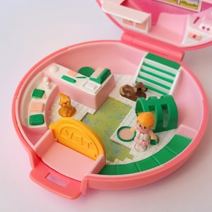 Puede incluir: Un juego de plástico rosa vintage, abierto para revelar una clínica veterinaria en miniatura. Incluye una pequeña figura, un perro marrón, un mostrador de recepción y otros accesorios. El juego es redondo y tiene una tapa con bisagras.