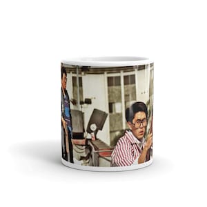 Pode incluir: Uma caneca de cerâmica branca com uma foto de duas pessoas. A pessoa à direita está vestindo uma camisa listrada vermelha e branca e óculos. A pessoa à esquerda está vestindo uma camisa listrada azul e branca. O fundo é uma imagem desfocada de um prédio.