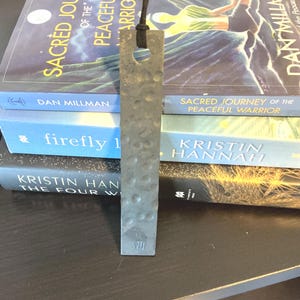 May include: A rectangular grey metal bookmark with a hammered texture and a circular hole at the top. A black cord is threaded through the hole. The Roman numeral VI is stamped near the bottom edge of the bookmark, which has a flat, matte finish.