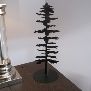 May include: A black metal tree sculpture with a circular base. The tree features a detailed silhouette of branches and needles. The sculpture is displayed on a wooden surface, with a lamp in the background.