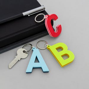 Può includere: Tre portachiavi a forma di lettera che rappresentano le lettere C, A e B in rosso, azzurro e verde lime. Ogni portachiavi ha un anello in metallo argentato. Una chiave argentata è attaccata al portachiavi A azzurro. È visibile anche una penna nera.