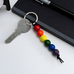 Könnte beinhalten: Ein regenbogenfarbener Perlen-Schlüsselanhänger mit einem silbernen Schlüssel. Der Schlüsselanhänger besteht aus Holzperlen und hat eine schwarze Schnur.