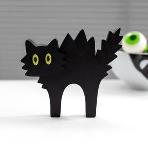 Puede incluir: Una figura de gato negro con la espalda arqueada y dentada y una cola puntiaguda. El gato tiene ojos grandes de color amarillo brillante y una silueta estilizada y angular. El diseño presenta bordes geométricos afilados en todo el cuerpo, la cabeza y el pelaje, creando una apariencia distintiva de perfil plano.