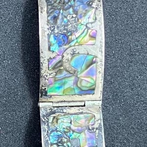 May include: A silver bracelet with rectangular links, each inlaid with iridescent abalone shell. The bracelet has a clasp and is displayed against a dark background. The abalone shell has blue, green, and purple hues.