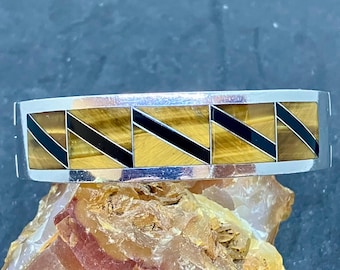 May include: A silver bracelet with a rectangular design. The bracelet features alternating sections of tiger's eye and black inlay. The bracelet is displayed on a rough, textured surface.