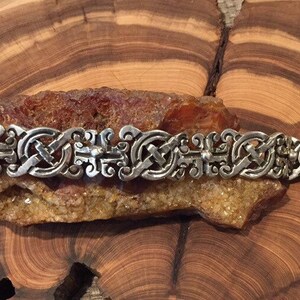 May include: A silver-toned metal bracelet featuring a repeating pattern of interconnected, ornate, scroll-like links. The design consists of symmetrical, curved elements and small spherical accents, creating a continuous, linked chain structure.