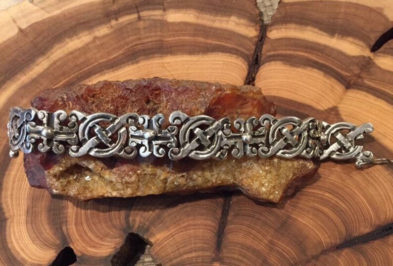 May include: A silver-toned metal bracelet featuring a repeating pattern of interconnected, ornate, scroll-like links. The design consists of symmetrical, curved elements and small spherical accents, creating a continuous, linked chain structure.