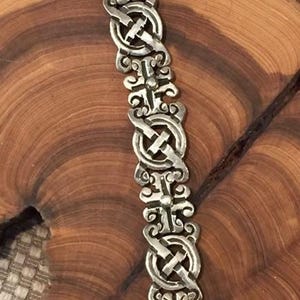 May include: A silver-toned metal bracelet featuring a repeating pattern of circular links connected by ornate, scroll-like cross pieces. The design consists of five identical segments with a metallic finish and a secure clasp mechanism at the top end.