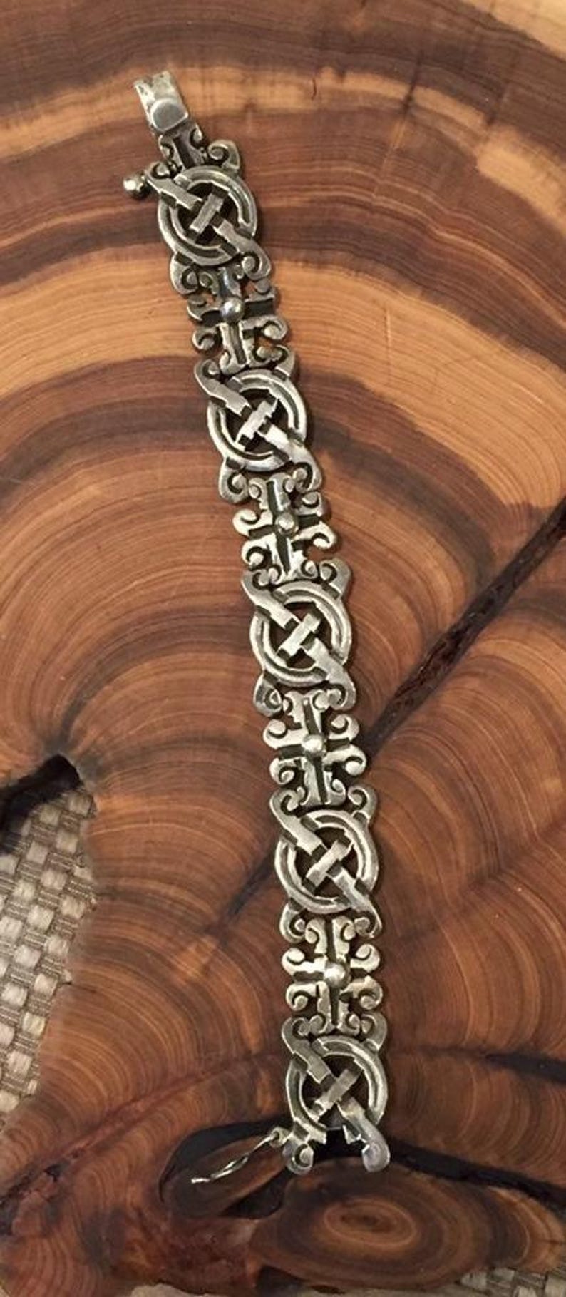 May include: A silver-toned metal bracelet featuring a repeating pattern of circular links connected by ornate, scroll-like cross pieces. The design consists of five identical segments with a metallic finish and a secure clasp mechanism at the top end.
