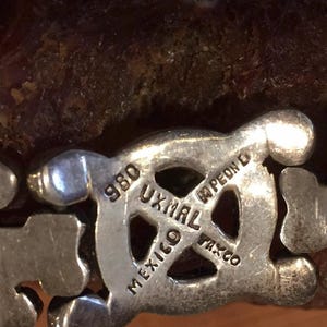 May include: A silver-toned metal link bracelet featuring a central circular emblem stamped with the text 980, UXMAL, MEXICO, TAXCO, and PEON D. The bracelet consists of repeating geometric shapes with a polished finish and visible hallmarks.