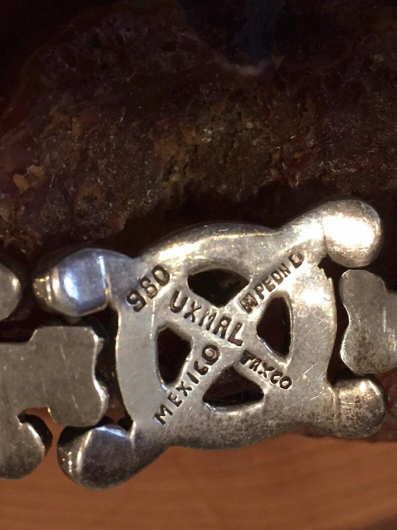 May include: A silver-toned metal link bracelet featuring a central circular emblem stamped with the text 980, UXMAL, MEXICO, TAXCO, and PEON D. The bracelet consists of repeating geometric shapes with a polished finish and visible hallmarks.