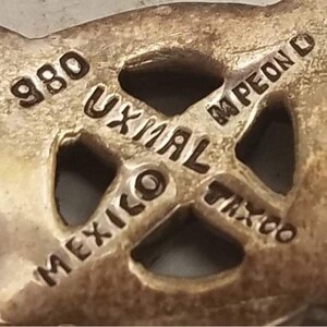 May include: A silver-toned metal jewelry piece featuring an openwork design with stamped text that reads 980, UXMAL, MPEON, MEXICO, and TAXCO. The surface has a metallic finish with visible indentations and raised lettering throughout the structure.