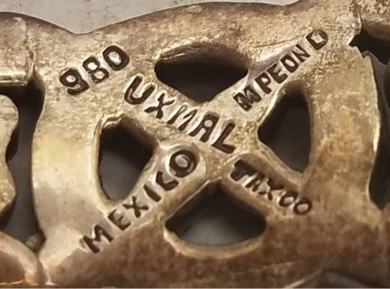 May include: A silver-toned metal jewelry piece featuring an openwork design with stamped text that reads 980, UXMAL, MPEON, MEXICO, and TAXCO. The surface has a metallic finish with visible indentations and raised lettering throughout the structure.