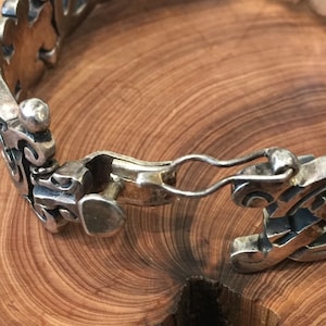 May include: A silver bracelet featuring a series of interconnected, ornate, openwork links with a polished metallic finish. The piece includes a secure hook-and-eye clasp mechanism that connects the two ends of the decorative, patterned metal band.
