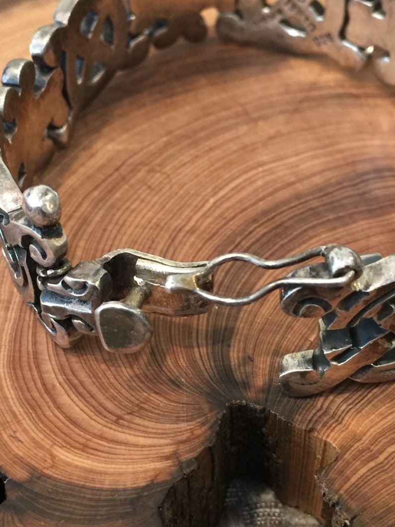 May include: A silver bracelet featuring a series of interconnected, ornate, openwork links with a polished metallic finish. The piece includes a secure hook-and-eye clasp mechanism that connects the two ends of the decorative, patterned metal band.