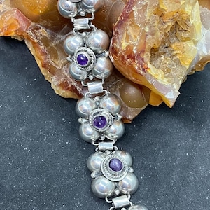 May include: A silver bracelet featuring a floral design with five clusters of silver spheres. Each cluster has a central, round, purple gemstone. The bracelet is displayed on a rough, textured, orange and brown stone surface.