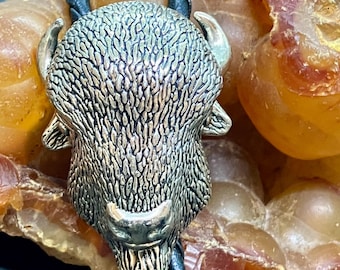 May include: A silver-toned bolo tie featuring a detailed bison head pendant. The bison has textured fur and small horns. The black leather cord is braided. The background is a mix of orange and brown.