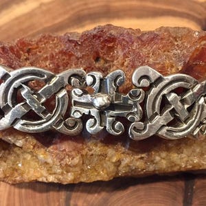 May include: A silver bracelet featuring a repeating pattern of interconnected circular links and a central rectangular link with a small raised sphere. The metal has a polished finish with dark recessed areas highlighting the intricate design details.