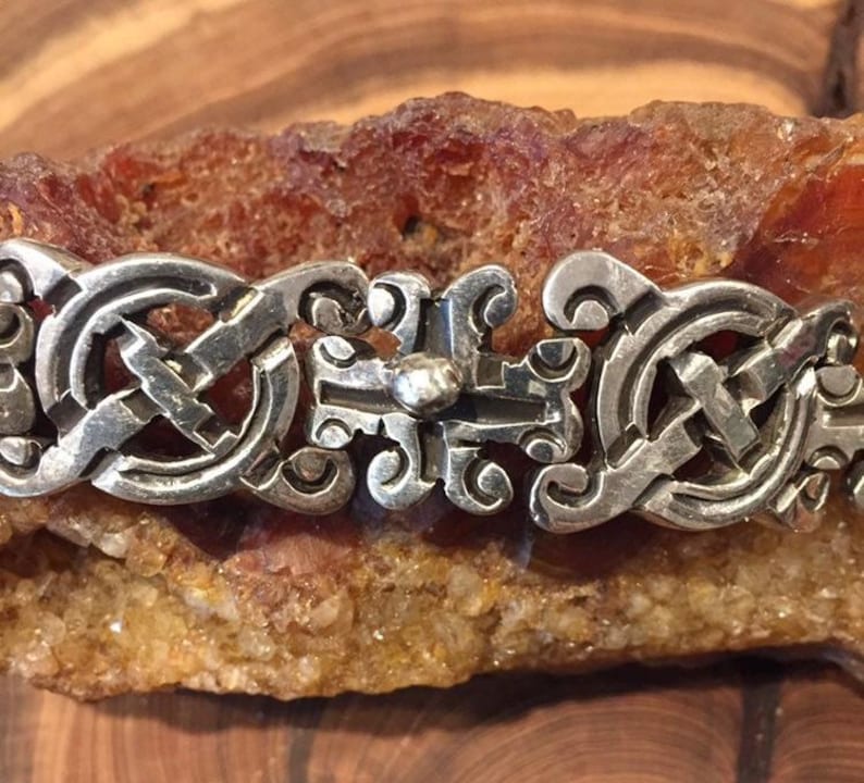 May include: A silver bracelet featuring a repeating pattern of interconnected circular links and a central rectangular link with a small raised sphere. The metal has a polished finish with dark recessed areas highlighting the intricate design details.