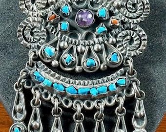 Puede incluir: Un colgante de plata con un diseño detallado que presenta piedras turquesa y amatista. El colgante tiene una franja de gotas de plata.