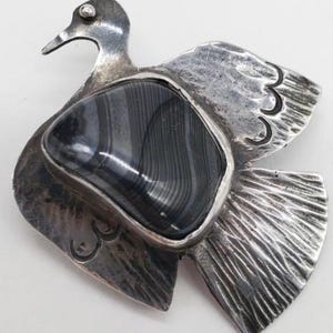 May include: A silver bird brooch with a black and white agate stone in the center. The bird has a detailed design and is a unique piece of jewelry.