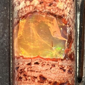 May include: A close-up of a small, iridescent opal gemstone set in a dark metal frame. The opal is a light orange color with flashes of green and blue.