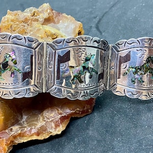 May include: A silver-toned bracelet with a series of rectangular panels, each featuring a horse design with abalone shell inlay. The bracelet has a scalloped edge and is displayed on a natural stone surface. The horse figures are in various poses.
