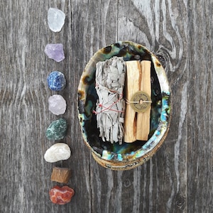 May include: A white sage smudge stick and a palo santo stick are bundled together and placed in a seashell on a wooden surface.  A row of colorful crystals are lined up on the left side of the image.