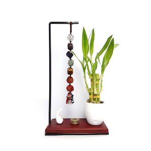 May include: A black metal stand with a red wooden base holds a lucky bamboo plant in a white pot. A string of seven colorful crystal beads hangs from the stand.