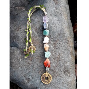 May include: A handmade gemstone pendant featuring a string of colorful, raw crystals in shades of red, orange, green, blue, and purple. The pendant is adorned with two small, round, bronze-colored coins and a decorative vine. The pendant is displayed on a gray rock.