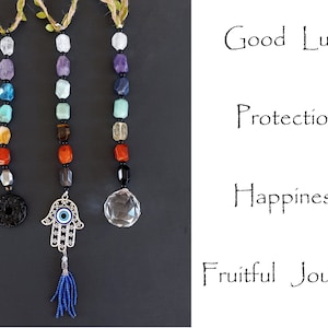 May include: Three crystal and gemstone beaded hanging charms with different charms at the bottom. The charms are a black stone circle, a silver Hamsa hand with a blue evil eye, and a clear crystal ball. The charms are labeled with the words "Good Luck", "Protection", "Happiness", and "Fruitful Journey".
