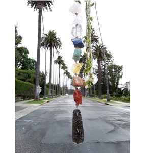 May include: A seven-chakra crystal necklace with a black obsidian pendant hanging from a string with green leaves. The necklace is hanging in front of a street lined with palm trees.