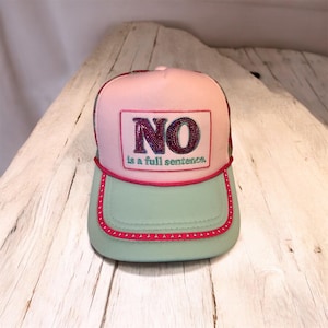 May include: A pink and green baseball cap with the word "NO" in sequined letters on the front. The text "is a full sentence" is below the word "NO". The cap has a pink brim and a green visor.