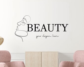 Può includere: Un design minimalista in bianco e nero di un viso di donna con la parola "BEAUTY" e il testo "your slogan here" sotto. Il design è su una parete bianca con due poltrone rosa e un tavolino da caffè in legno di fronte.